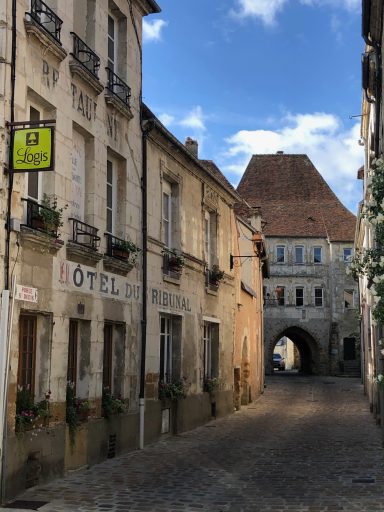Rue pavée avec des bâtiments en pierre, balcons fleuris et un arc au loin.
