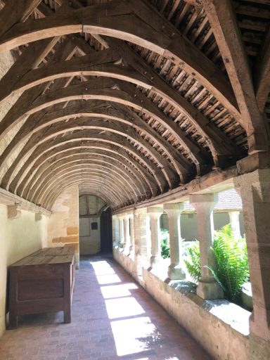 Couloir avec voûte en bois, colonnes en pierre, lumière naturelle et verdure à l'extérieur.